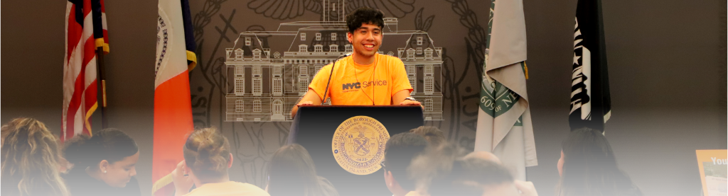 Young man wearing an orange NYC Service shirt stands behind an Office of the Borough President of Staten Island podium against a wall with a white outline of the Staten Island Borough Hall and flags on both sides of the young man. Young people are pictured toward the bottom of the picture as they face the young man at the podium.